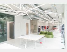 Heller, moderner Raum mit offenen Decken, sichtbaren Lüftungselementen und linear angeordneten LED-Leuchten. Der Raum umfasst weiße Tische, grüne modulare Sitzmöbel, Kleiderständer mit Kleidung sowie eine Tischtennisplatte im Hintergrund.