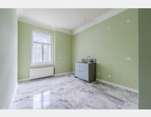 Leerer Raum mit hellgrünen Wänden, dekorativer Stuckleiste an der Decke und glänzendem, marmoriertem Fliesenboden. Links ein großes Fenster mit Heizkörper darunter, rechts ein kleiner Küchenunterschrank mit Spüle und mehreren Steckdosen an der Wand.