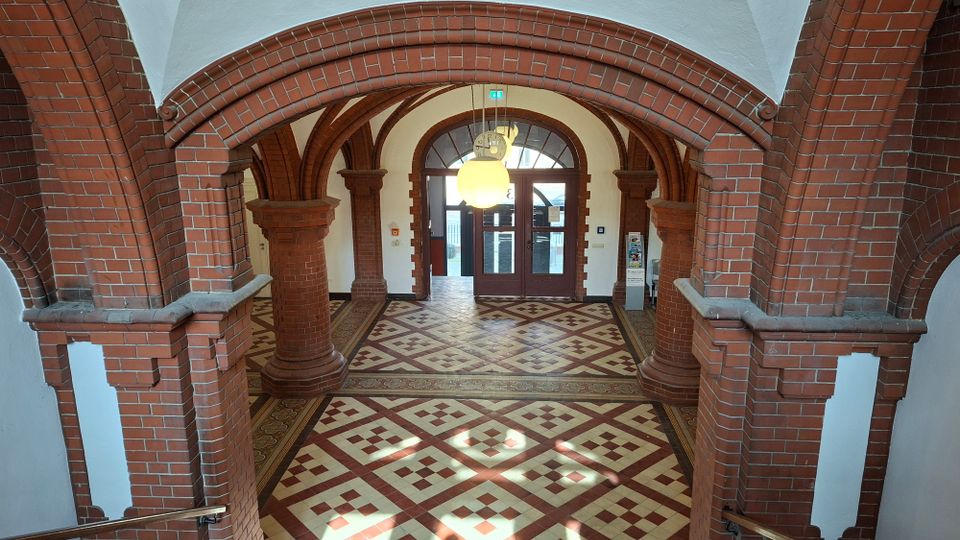 Eingangshalle eines historischen Backsteingebäudes mit Rundbögen und massiven Pfeilern, weiß verputztem Gewölbe und großem Kugelleuchter. Der Boden ist mit geometrisch gemusterten Fliesen ausgelegt und führt zu einer Doppeltür mit Oberlichtfenster.
