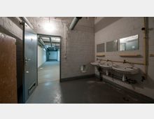 Ein einfacher Waschraum mit drei weißen Waschbecken an einer weißen Ziegelwand, darüber befinden sich drei rechteckige Spiegel. Links führt eine geöffnete Metalltür in einen kargen Flur mit Betonboden und sichtbaren Rohren an der Decke.