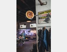 Innenraum einer Motorradwerkstatt mit einer hängenden Neon-Leuchtreklame, die einen Löwen zeigt, sowie einem Poster an der Wand. Im Hintergrund sind Werkbänke, Hängelampen, Regale und ein Motorrad sichtbar, während im Vordergrund Motorradkleidung an einer Kleiderstange hängt.