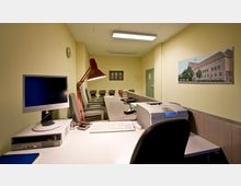 Ein schlichtes Büro mit gelben Wänden, einem langen Schreibtisch, Computermonitor und Drucker im Vordergrund, sowie einer roten Schreibtischlampe. Im Hintergrund befinden sich mehrere Stühle entlang der Wand, ein Kleiderständer, sowie ein geschlossenes Tür- und Fenstermotiv und ein an der Wand hängendes Foto eines Gebäudes.
