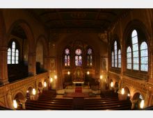Innenansicht einer historischen Kirche mit Backsteinmauern, Spitzbogenfenstern mit buntem Glas und verzierten Wandmalereien. Im Vordergrund sind Holzbänke, im Hintergrund der Altar mit leuchtenden Glasfenstern und einer roten Teppichführung sichtbar.