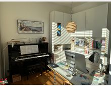 Ein heller Arbeitsraum mit einem schwarzen Klavier auf der linken Seite, auf dem Notenblätter liegen, und einem Schreibtisch mit einem Computerbildschirm in der Mitte. Im Hintergrund befindet sich ein großer weißer Einbauschrank mit offenen und geschlossenen Fächern, die Bücher und Büromaterial enthalten. Eine geflochtene Hängelampe hängt von der Decke.