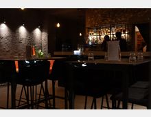 Dunkle, moderne Bar mit hohen Tischgruppen und schwarzen Stühlen, beleuchtet durch Pendelleuchten. Im Hintergrund eine Steinwand mit Regal, das mit Flaschen bestückt ist, und eine Theke mit Personen.