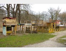 Holzspielplatz mit improvisierten Spielstrukturen, darunter ein kastenförmiges Haus mit Treppe und Balken sowie eine Konstruktion in Form eines Schiffes, umgeben von kahlen Bäumen und Grasflächen. Im Hintergrund sind Gebäude und weitere Spielgeräte zu erkennen.