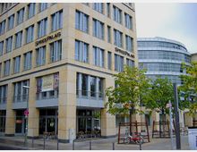 Eckansicht eines modernen Bürogebäudes mit der Aufschrift "Spreepalais" an der Fassade. Das Gebäude besteht aus beigen Fassadenplatten, großen Fenstern und einem angrenzenden Bereich mit Bäumen, Sitzgelegenheiten und einem Fahrradständer.