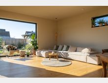 Helles Wohnzimmer mit Parkettboden, einem großen Ecksofa in Beige, einem runden Teppich sowie einem kleinen Holztisch. Große Fensterfronten lassen viel Tageslicht herein und geben den Blick auf einen Garten frei; Pflanzen und Dekoration ergänzen den Raum.