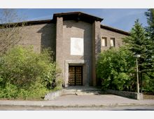 Backsteinbau mit einem breiten Eingangsportal aus Holz, flankiert von Betonrahmen und umgeben von dichtem Grün, darunter Bäume und Büsche. Der Gehweg davor ist uneben, und rechts steht ein schlichter Straßenlampenmast.