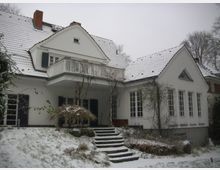Schneebedecktes Einfamilienhaus mit weißer Putzfassade, Satteldach und einem kleinen Balkon mit weißem Geländer; im Vordergrund eine verschneite Treppe und kahles Rankwerk an der Hauswand.
