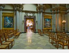 Innenraum einer historischen Kirche mit verzierten Säulen, Wandmalereien in vergoldeten Rahmen und Holzstühlen, die in Reihen angeordnet sind. Ein kunstvoll gestalteter Türrahmen aus Marmor führt in einen weiteren, hell erleuchteten Bereich mit einer prunkvollen Deckenkonstruktion.