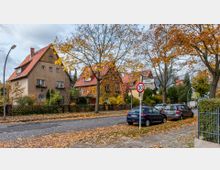 Straßenszene in einem Wohngebiet mit Einfamilienhäusern, die rote Ziegeldächer und beige Fassaden haben. Die Bäume tragen herbstlich gefärbtes Laub, am Straßenrand sind geparkte Autos sowie ein Tempo-30-Verkehrsschild zu sehen.