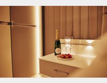 Raum mit indirekter Beleuchtung, einer Holzvertäfelung und einem weißen Möbelstück, auf dem eine Flasche Champagner, zwei leere Sektgläser und ein Teller mit Erdbeeren stehen.