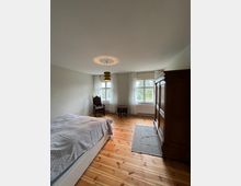 Schlafzimmer mit Holzboden, einem Bett auf der linken Seite, einem antiken Stuhl und einem kleinen Tisch vor zwei Fenstern mit weißen Vorhängen. Rechts steht ein großer Holzschrank, und eine schlichte Deckenlampe hängt in der Mitte des Raumes.