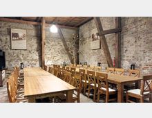 Ein rustikaler Innenraum mit langen Holztischen und passenden Stühlen, umgeben von Wänden aus unverputztem Ziegelstein. Freiliegende Holzbalken an der Decke und dekorative Laternen auf den Tischen betonen den ländlichen Stil, während an den Wänden Bilder aufgehängt sind.