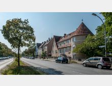 Straßenszene mit mehrstöckigen Wohnhäusern im klassischen Stil auf der rechten Seite, teilweise mit roten Ziegeldächern und Gauben. Links verläuft eine zweispurige Fahrbahn mit parkenden Autos und Bäumen entlang der Mittelinsel, unter klarem Himmel.