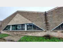 Ein modernes Gebäude aus Ziegelsteinen mit schräg verlaufendem Mauerwerk und großen, trapezförmigen Glasfenstern. Vor dem Gebäude liegt eine grasbedeckte Fläche und ein betonierter Gehweg.