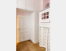 Ein heller Innenraum mit weißer Tür, hohen Einbauschränken und einem Holzfußboden in Fischgrätmuster. Rechts im Bild ist eine Spielhütte mit Fenster und einem Vorhang mit bunten Punkten zu sehen.