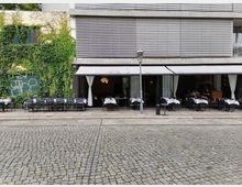 Straßencafé mit außen aufgestellten Tischen und Stühlen, teilweise überdacht durch eine Markise. Die Hausfassade besteht aus Beton und Glas, teils mit Begrünung; im Vordergrund Kopfsteinpflaster.
