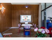 Konferenzraum mit Holzwänden, einem langen Tisch, umgeben von Stühlen mit farbigen Polstern, und einem Bildschirm an der Wand, der den Text "Change is a Team Sport" zeigt. Im Vordergrund stehen Blumen in einer Vase, und rechts befindet sich eine Glastrennwand mit Vorhängen.