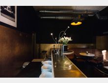 Eine dimm beleuchtete Bar mit Metalltheke, darauf weiße Schüsseln, Blumenvasen und moderne Stehlampen. Im Hintergrund sind Sitznischen mit runden Tischen und braunen Lederbänken sowie eine Decke mit sichtbaren Lüftungsrohren zu sehen.