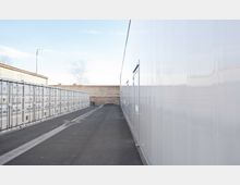 Ein langer, asphaltierter Weg verläuft zwischen einer Reihe weißer Lagercontainer auf der linken Seite und einer großen, glänzenden Wand auf der rechten Seite. Im Hintergrund sind eine Backsteinmauer und ein kleiner Baum zu sehen.
