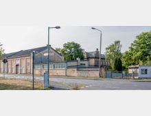 Straßenszene mit historischen Gebäuden aus Backstein, darunter ein großes Lagerhaus mit Flachdach und ein mehrstöckiges Gebäude mit verziertem Dach first, in einem teilweise verwahrlosten Zustand. Im Vordergrund sind Straßenschilder, Laternen und ein Gehweg zu sehen, im Hintergrund befinden sich Bäume.