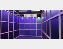 Ein Raum mit Wänden aus einem Holzrahmen-Gittersystem, gefüllt mit dunkelblauen Paneelen, beleuchtet durch lila und weiße LED-Lichter. An der Decke befinden sich leuchtende Hinweise mit Text und QR-Codes.
