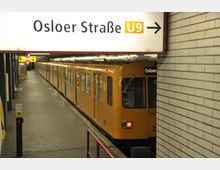 Ein U-Bahnhof mit gelber U-Bahn der Linie U9, die an der Station "Osloer Straße" hält. Im Vordergrund ein Schild mit der Aufschrift "Osloer Straße U9" und einem Pfeil, umgeben von weiß gefliesten Wänden und einer Bahnsteigkante.