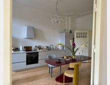 Moderne Küche mit weißer Einbauzeile, Edelstahlspüle, Dunstabzugshaube und Ofen, ergänzt durch Dekorationen und Küchenutensilien auf der Arbeitsplatte. Im Vordergrund ein roter Esstisch mit einer Vase voller Blumen und gelben, gepolsterten Stühlen; Fischgrätparkettboden und weiße Türen im Hintergrund.