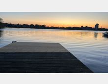 Ein ruhiger See bei Sonnenuntergang mit einem hölzernen Steg im Vordergrund. Am Horizont sind silhouettierte Gebäude, Bäume und ein einzelner hoher Turm zu sehen.