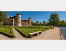 Large historic palace with cream-colored walls and red roofs, featuring two round corner towers, viewed across formal hedged garden paths. A sandy walkway runs along a lake on the right, lined with trees and a bench in the foreground.