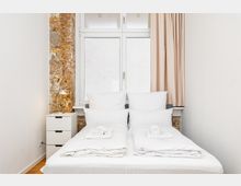 Schlichtes Schlafzimmer mit zwei zusammengestellten Einzelbetten in weißer Bettwäsche vor einem großen Fenster mit Vorhang. Links stehen ein kleiner weißer Nachttisch mit Lampe und eine freigelegte Backsteinwand als Akzent neben weißen Wänden.