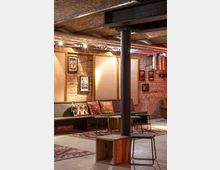 Ein gemütlicher Raum mit Backsteinwänden, Decken aus Sichtbeton und sichtbaren Rohren. Der Raum ist ausgestattet mit einer langen Sitzbank, bunten Kissen, kleinen Tischen und Hockern sowie Teppichen mit geometrischen Mustern.