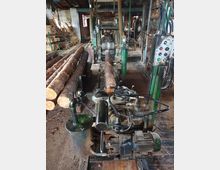 Eine Holzwerkstatt mit einer Sägeanlage, die Baumstämme bearbeitet. Im Vordergrund befindet sich eine Maschine mit Motor und Steuerungselementen, während auf der linken Seite mehrere entrindete Baumstämme auf Holzbalken liegen.