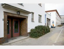 Wohngebäude in moderner Architektur mit weißer Fassade und braunem Eingangsbereich, bestehend aus zwei Türen mit blau umrandeten Glaseinsätzen; davor ein kleiner gepflasterter Gehweg mit angrenzenden Sträuchern. Im Hintergrund sind weitere Gebäude und eine Straße zu sehen.