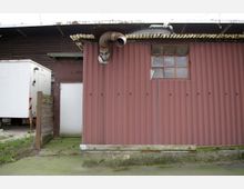 Außenansicht eines einfachen Gebäudes mit Wellblechwänden in rötlicher Farbe, einem kleinen Fenster mit Holzrahmen und einem herausragenden Abzugsrohr. Daneben ist ein weißer, rechteckiger Anhänger und ein Holzzaun sichtbar.