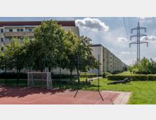 Ein kleiner Sportplatz mit rotem Bodenbelag und einem Metalltor im Vordergrund. Im Hintergrund sind Apartmentgebäude, Bäume, Grünflächen und ein Strommast zu sehen.