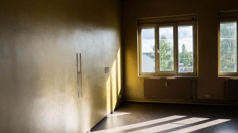 Leerer Innenraum mit beigefarbenen Wänden und dunklem Boden, von zwei großen Fenstern durch Tageslicht stark ausgeleuchtet. Unter den Fenstern befinden sich Heizkörper, und durch die Scheiben sind Bäume und Gebäude im Außenbereich zu sehen.