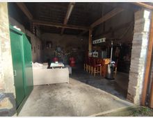 Ein einfacher Innenraum eines umgebauten Gebäudes mit rustikalen Wänden, Holzbalken an der Decke und offener Struktur. Der Raum enthält eine Sitzgruppe mit Sofa, einen Tresen mit Barhockern und Dekorationselemente wie Schilder und Lichterketten an den Wänden.