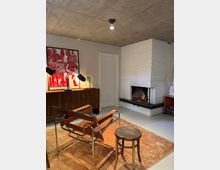 Modern living room with an exposed concrete ceiling, a white-painted brick corner fireplace with a fire burning, and a minimalist seating area on a tan rug. A low wooden sideboard with desk lamps and a large red poster sits against a gray wall beside a white door.