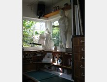Arbeitszimmer mit großem Fenster und Blick auf einen Garten. Auf der breiten Fensterbank stehen zwei weiße Statuen, darunter eine große griechische Figur; der Raum verfügt über einen Arbeitsplatz mit Schreibtisch, Regalen mit Materialien und einer Holzkommode.