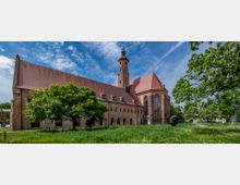 Backsteingebäude mit gotischen Elementen, darunter große Spitzbogenfenster und ein hoher Kirchturm, umgeben von grüner Wiese und Bäumen unter blauem Himmel. Im Vordergrund sind ein großer Baum und eine Skulptur zu sehen.