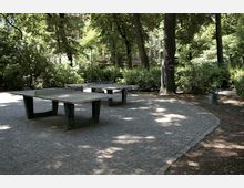 Spielplatzbereich in einem Park mit Tischtennisplatten auf einer gepflasterten Fläche, umgeben von hohen Bäumen und dichtem Grün. Im Hintergrund sind Gebäude und Bänke sichtbar.