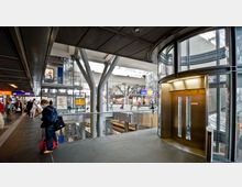 Großes, modernes Bahnhofsgebäude mit mehreren Ebenen, offenen Stahlträgern und viel Glas. Im Vordergrund ein gläserner Aufzug, dahinter Geschäfte, Rolltreppen und Bahnsteige, durchzogen von Passanten.