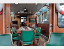 Innenraum eines klassischen Bootes mit Holzwänden und -decke, einem großen zentralen Esstisch mit grünen Lederstühlen und dekoriert mit Geschirr und einer Vase mit Blumen. An den Seiten befinden sich Fenster mit Vorhängen, die den Blick nach draußen ermöglichen.