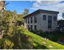 Zweistöckiges Gebäude aus hellem Backstein mit modernen Fenstern, umgeben von einem gepflegten Garten mit Rasen, Bäumen und Pflanzen. Im Vordergrund führen Steinplatten durch das Grün, im Hintergrund ist blauer Himmel erkennbar.