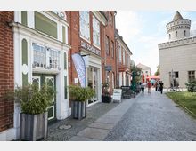 Gepflasterte Fußgängerstraße mit roten Backsteingebäuden, in denen Geschäfte wie "Das schöne Bett" untergebracht sind. Auf der rechten Seite ein Gebäude mit Zinnen und ein Turm, daneben Gehwege und Straßencafés mit Sonnenschirmen; im Hintergrund Passanten und Fahrräder.