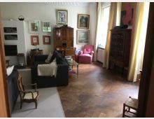 Wohnzimmer mit Holzboden, ausgestattet mit dunkelgrauen Sofas, einem roten Sessel, einem antiken Holzschrank und einer Vitrine. An den Wänden hängen zahlreiche gerahmte Kunstwerke, und durch große Fenster fällt Tageslicht, ergänzt durch lange Vorhänge in gelblichen und rötlichen Tönen.