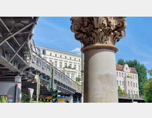 Detailaufnahme einer verzierten Steinsäule im Vordergrund, daneben eine erhöhte Stahlkonstruktion einer Hochbahntrasse. Im Hintergrund sind klassizistische Stadtgebäude mit hellen Fassaden sowie Bäume und Verkehrsampeln zu sehen.